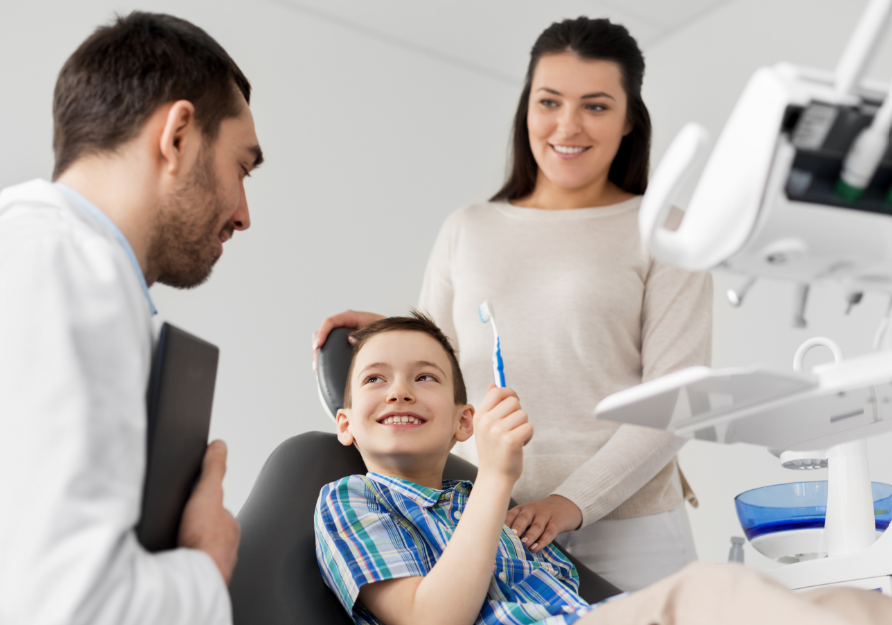 Little boy in dental chair holding toothbrush while mom and dentist smile